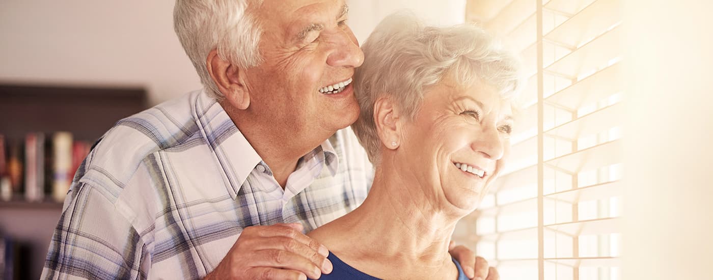 A happy older man and woman standing together, looking through window blinds.